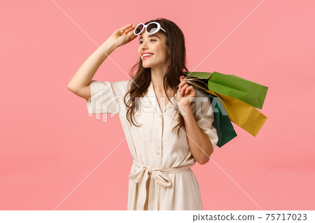 Carefree enthusiastic woman in dress, taking off glasses and looking happily sideways as enjoying beautiful day, fantastic shopping with discounts, holding shop bags behind back, pink background 75717023