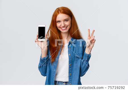 Cute and cheerful alluring redhead female in denim shirt, showing peace gesture smiling and holding smartphone, show app, company website or shopping internet store on mobile screen, white background Cute and cheerful alluring redhead female in denim shirt, showing peace gesture smiling and holding smartphone, show app, company website or shopping internet store on mobile screen, white background 75719292