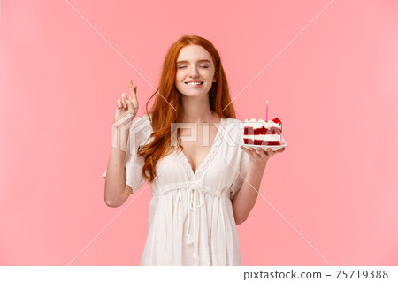 Celebration, happiness concept. Attractive dreamy redhead girl in lovely white dress, holding b-day cake, cross finger good luck, bite lip tempting close eyes and making wish to blow candle 75719388