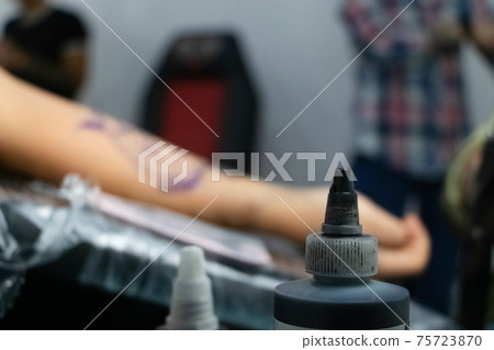 Tattoo concept. A jar of paint for tattoos, stands against the background of a man's hand. You can also see how the master fills the pattern on the forearm. Tattoo concept. A jar of paint for tattoos, stands against the background of a man's hand. You can also see how the master fills the pattern on the forearm. 75723870