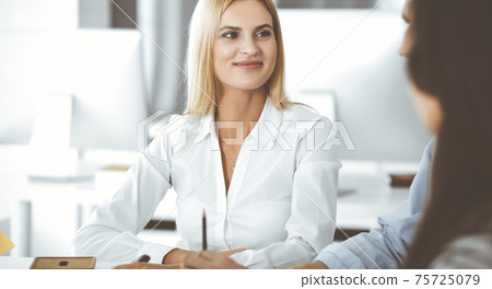 Group of business people discussing questions at meeting. Headshot of blonde businesswoman while smiling to her colleague at office negotiation. Teamwork and cooperation in corporate occupation Group of business people discussing questions at meeting. Headshot of blonde businesswoman while smiling to her colleague at office negotiation. Teamwork and cooperation in corporate occupation 75725079