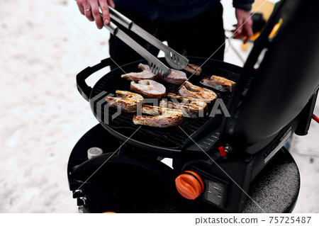 Man grilling delicious fish on a portable BBQ, Snowy winter barbecue outdoors in the cold . Man grilling delicious fish on a portable BBQ, Snowy winter barbecue outdoors in the cold . 75725487