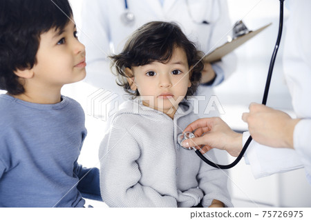 Woman-doctor examining a child patient by stethoscope. Cute arab toddler and his brother at physician appointment. Medicine concept Woman-doctor examining a child patient by stethoscope. Cute arab toddler and his brother at physician appointment. Medicine concept 75726975