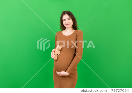 Happy pregnant woman holding a teddy bear against her belly at Colored background. Young mother is expecting a baby. Copy space Happy pregnant woman holding a teddy bear against her belly at Colored background. Young mother is expecting a baby. Copy space 75727064