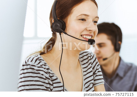 Casual dressed young woman using headset and computer while talking with customers online. Group of operators at work. Call center, business concept Casual dressed young woman using headset and computer while talking with customers online. Group of operators at work. Call center, business concept 75727115
