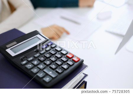 Calculator and binders with papers are waiting to be processed by business woman or bookkeeper working at the desk in office back in blur. Internal Audit and tax concept 75728305