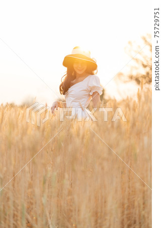Happy young beautiful woman wearing black hat and white dress enjoying herself walking in the golden barley filed on a late afternoon Happy young beautiful woman wearing black hat and white dress enjoying herself walking in the golden barley filed on a late afternoon 75729751