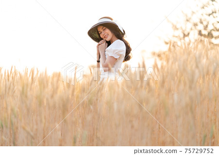 Happy young beautiful woman wearing black hat and white dress enjoying herself daydreaming in the golden barley filed on a late afternoon, 75729752