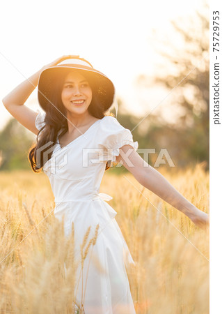 Happy young beautiful woman wearing black hat and white dress enjoying herself walking in the golden barley filed on a late afternoon Happy young beautiful woman wearing black hat and white dress enjoying herself walking in the golden barley filed on a late afternoon 75729753