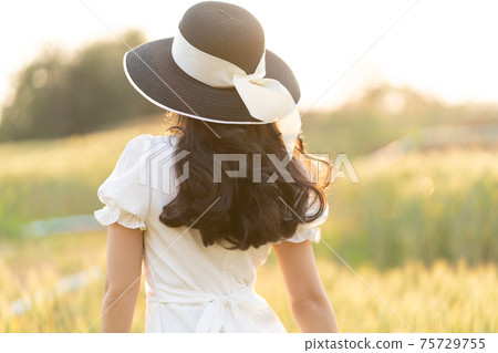 Back view of young woman wearing black hat and white dress walking alone at a barley field on a late afternoon before sunset Back view of young woman wearing black hat and white dress walking alone at a barley field on a late afternoon before sunset 75729755