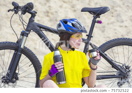 Beautiful girl cyclist in yellow with tea and thermos in hand on the background of sand 75732522