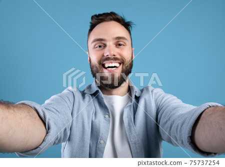Joyful bearded guy taking selfie on blue studio background 75737254