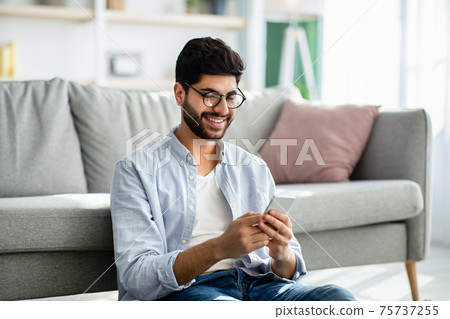 Arab guy texting and browsing internet on smartphone, resting at home, leaning on sofa 75737255