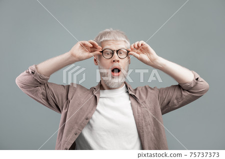 Surprised albino blond man touching glasses with open mouth, guy posing over grey studio background 75737373