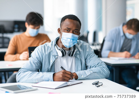 Male black student in mask sitting at desk and writing Male black student in mask sitting at desk and writing 75737389