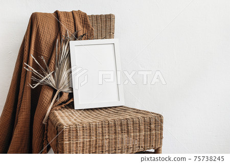 Exotic tropical boho interior still life. Blank white vertical picture frame mock-up on rattan chair. Cinnamon muslin plaid with dry palm leaf. White wall background. Empty copy space, no people. Exotic tropical boho interior still life. Blank white vertical picture frame mock-up on rattan chair. Cinnamon muslin plaid with dry palm leaf. White wall background. Empty copy space, no people. 75738245