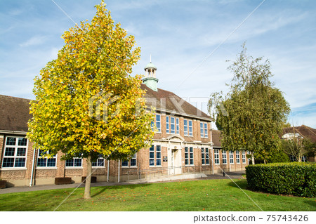 A historic brick school building surrounded by trees where the autumn leaves have begun under the blue sky in the suburbs of London 75743426