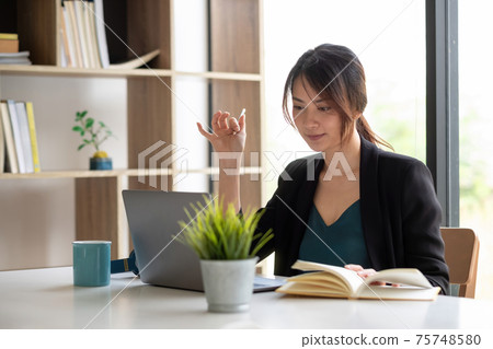 Asian businesswoman talking to colleague team in video call conference writing note on book with smile face Asian businesswoman talking to colleague team in video call conference writing note on book with smile face 75748580