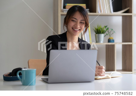 Happy Asian woman concentrated working video conference team with laptop computer and writing on notepad. Business Team Online Happy Asian woman concentrated working video conference team with laptop computer and writing on notepad. Business Team Online 75748582