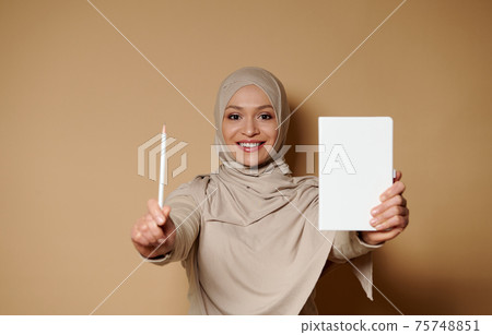 Charming arabian Muslim woman with beautiful smile holding a white diary and pencil. 75748851