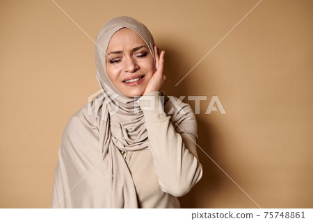 Muslim woman in a strict formal traditional dress with a head covered with a hijab suffers from a toothache. Beige background with copy space Muslim woman in a strict formal traditional dress with a head covered with a hijab suffers from a toothache. Beige background with copy space 75748861
