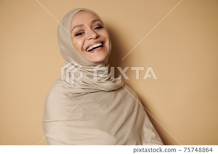 Attractive woman in strict religious clothes and head covered in hijab, laughs looking at camera against beige background with copy space 75748864