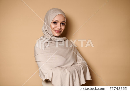 Attractive woman in strict religious clothes and head covered in hijab, cute smiling looking at camera against beige background with copy space 75748865