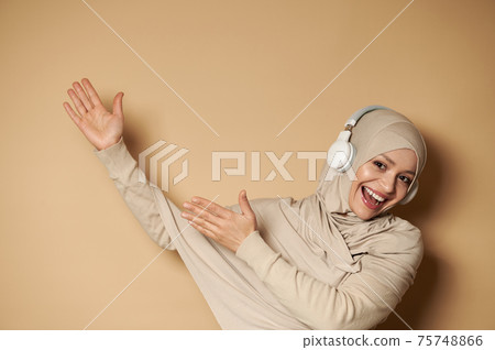 Gorgeous Muslim woman in hijab listening to the music in headphones, pointing hands on beige background with copy space Gorgeous Muslim woman in hijab listening to the music in headphones, pointing hands on beige background with copy space 75748866