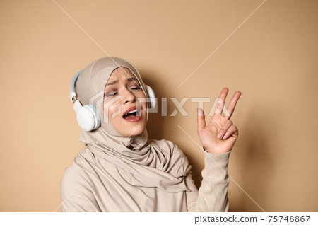 Gorgeous Muslim woman in hijab listening to the music in headphones, singing and dancing on beige background 75748867