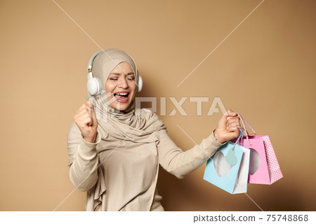 Muslim woman in hijab with headphones holding colored paper bags in her hands enjoying the upcoming religious holidays Muslim woman in hijab with headphones holding colored paper bags in her hands enjoying the upcoming religious holidays 75748868