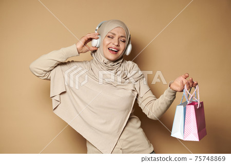 Beautiful Muslim woman in hijab listening to the music and holding colored gift bags. Copy space 75748869