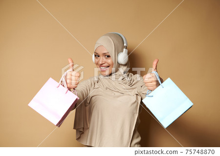 Attractive woman in hijab and headphones holding pink and blue gift paper bags in the hands and showing thumbs up, expressing happiness of upcoming religious holidays 75748870