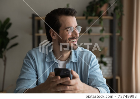 Close up smiling dreamy man wearing glasses distracted from phone Close up smiling dreamy man wearing glasses distracted from phone 75749998