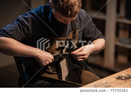 a young man is engaged in the family craft of making leather shoes in a workshop. 75750362