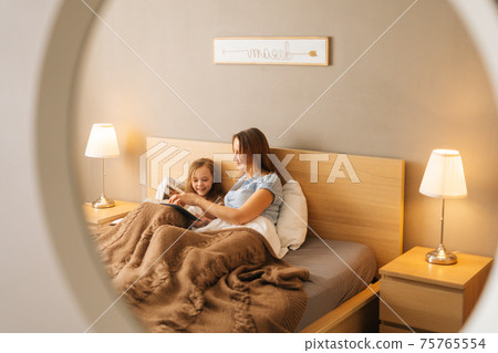 Reflection in mirror of young happy mother and adorable cheerful daughter reading children book together lying in bed before going to sleep. 75765554