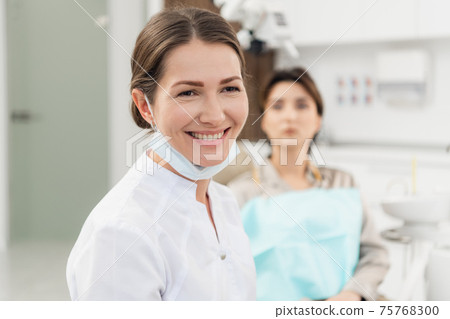 A portrait of a smiling dentist in her office, with a patient in A portrait of a smiling dentist in her office, with a patient in 75768300