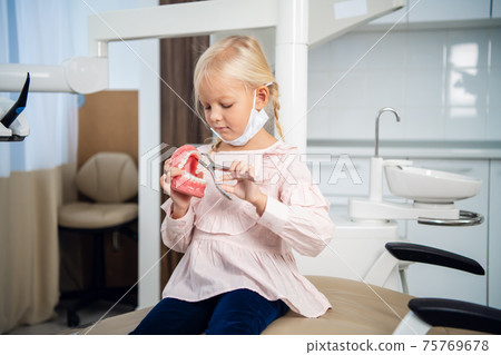 A sweet little girl playing with an artificial jaw and medical instruments in a dental clinic 75769678