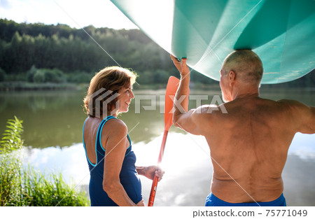 Rear view of senior couple carrying paddleboard by lake in summer. Rear view of senior couple carrying paddleboard by lake in summer. 75771049