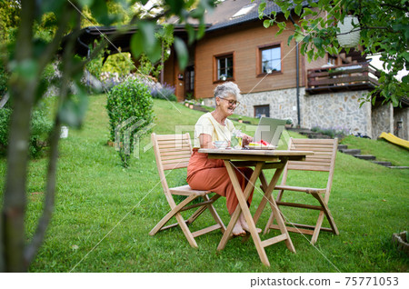 Happy senior woman with laptop working outdoors in garden, home office concept. 75771053