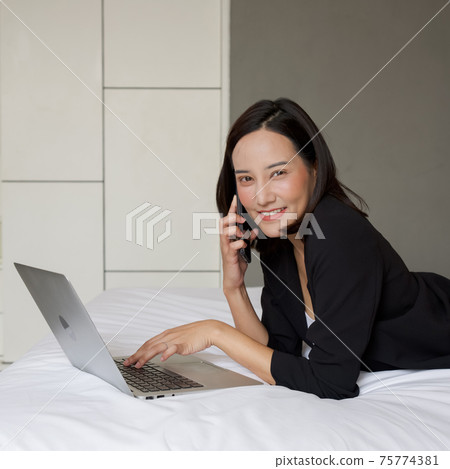 An asian woman in black suit use the laptop computer and mobile phone to work in the morning in bed. 75774381