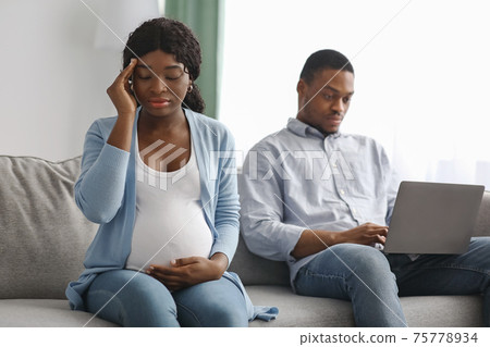 Upset pregnant african woman sitting by her husband using laptop 75778934