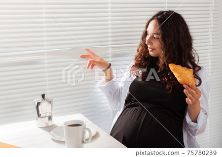 Happy pregnant young beautiful woman eating croissant and looking througt blinds during morning breakfast. Concept of pleasant morning and positive attitude during pregnancy 75780239
