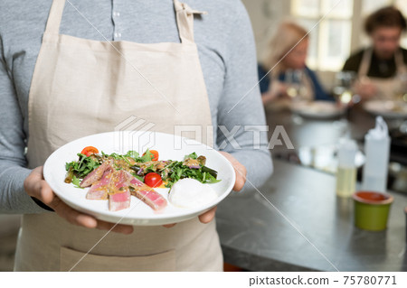 Midsection of mature man in pullover and apron holding plate with selfmade vegetable salad 75780771