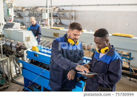 Two young intercultural male workers of modern plant discussing online technical data in workshop 75780994