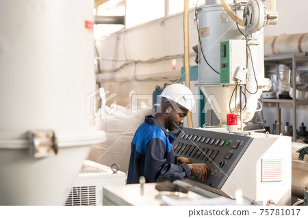 African worker standing in front of control panel of huge industrial machine and adjusting mechanism 75781017