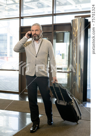 Mature businessman with smartphone by ear pulling suitcase while entering hall of hotel Mature businessman with smartphone by ear pulling suitcase while entering hall of hotel 75781324
