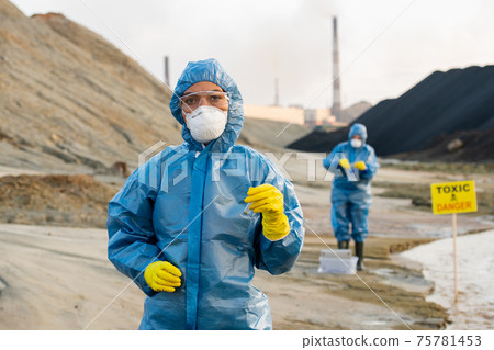Young serious female researcher in protective workwear holding sample of toxic soil in flask 75781453