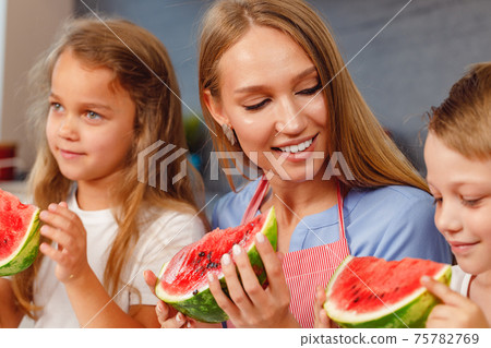 Woman with her daughter and son eating watermelon in kitchen 75782769