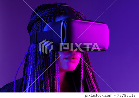 Young woman with afro braids using VR glasses on dark purple neon background 75783112