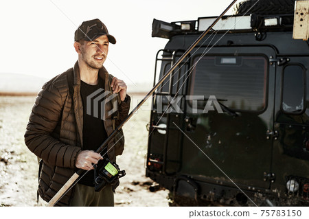 Young fisherman standing near his car and holding fishing equipment Young fisherman standing near his car and holding fishing equipment 75783150